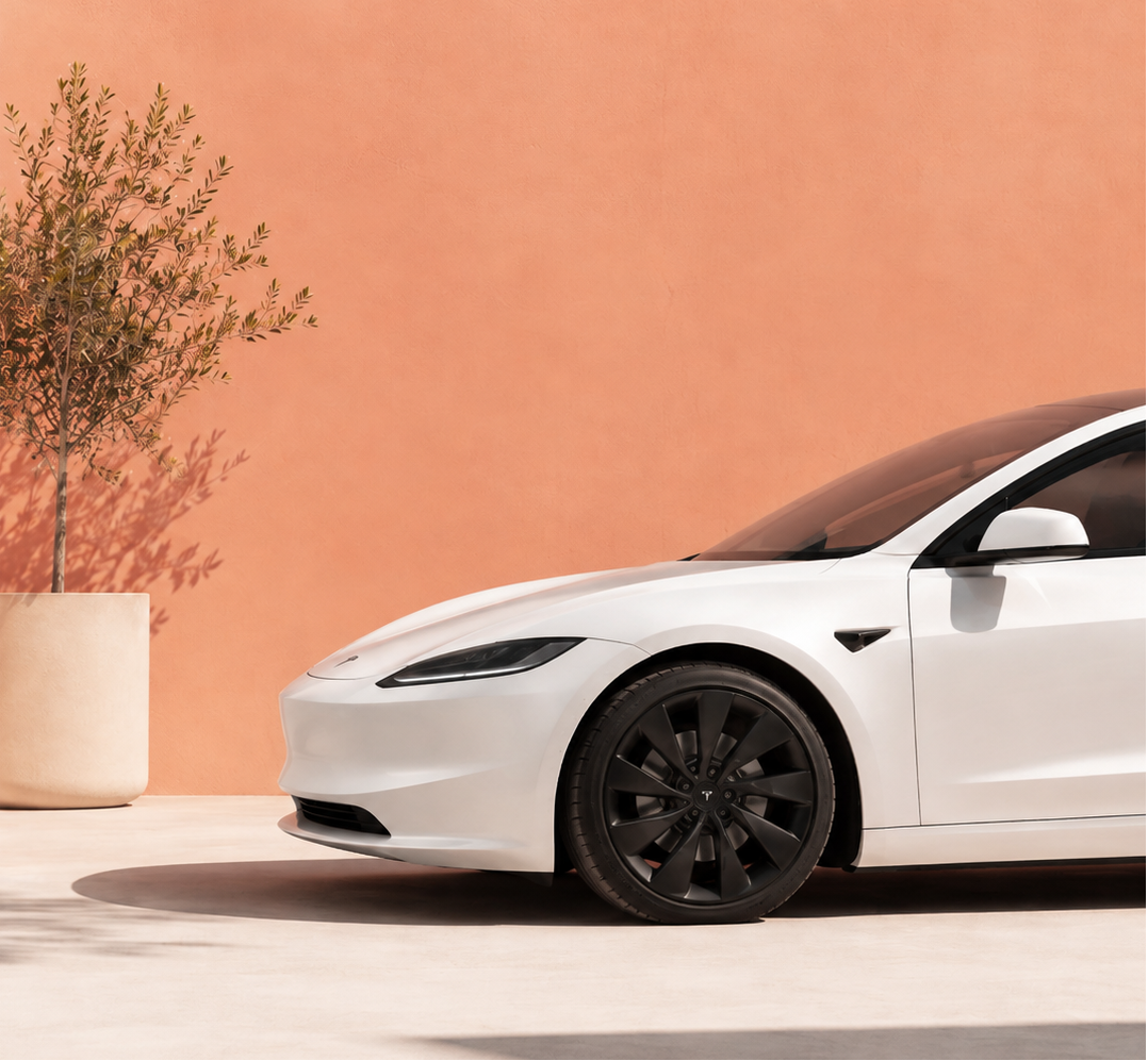 A white Tesla Model S shown from a side-front angle, parked outdoors against a warm terracotta wall with a potted tree and a light-colored column visible.