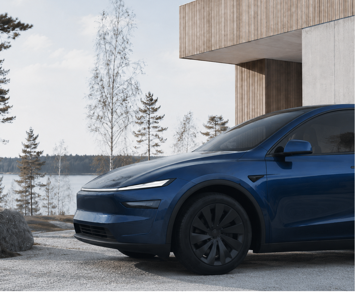 A dark blue Tesla Model Y SUV parked outdoors on a gravel surface next to a modern wooden building, with trees and water in the background.